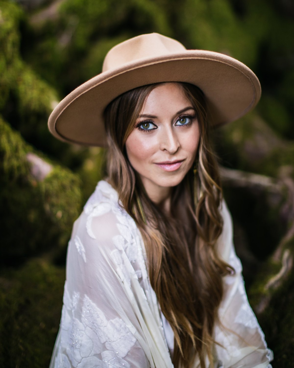 Portrait of Leanne in a tan wide-brimmed hat and white lace shawl, smiling softly against mossy rocks