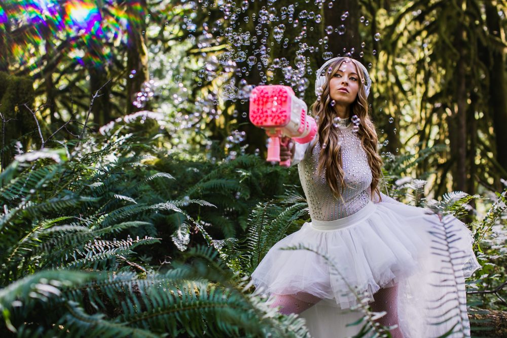 Leanne in white headphones and a crystal-studded bodysuit firing a pink bubble gun in the forest
