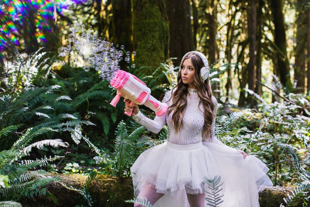 Leanne in white headphones and a tulle skirt holding a pink bubble gun among ferns