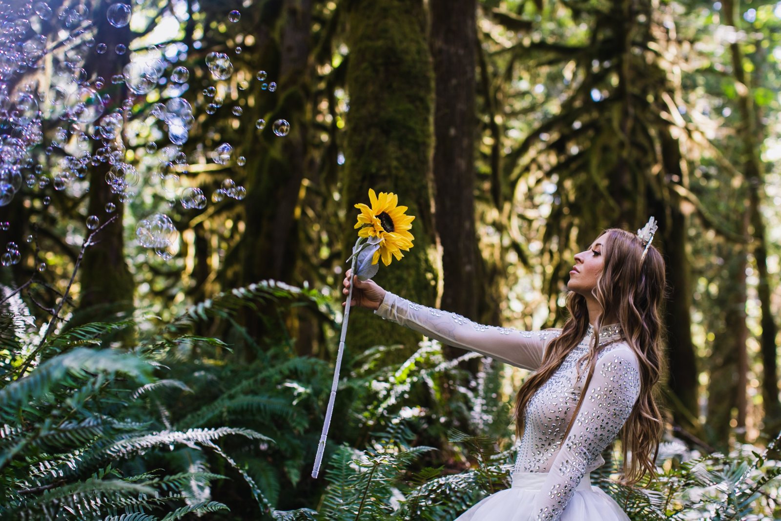 Leanne in a crystal-studded white bodysuit and crown holding a sunflower out toward a cloud of bubbles in a fern forest