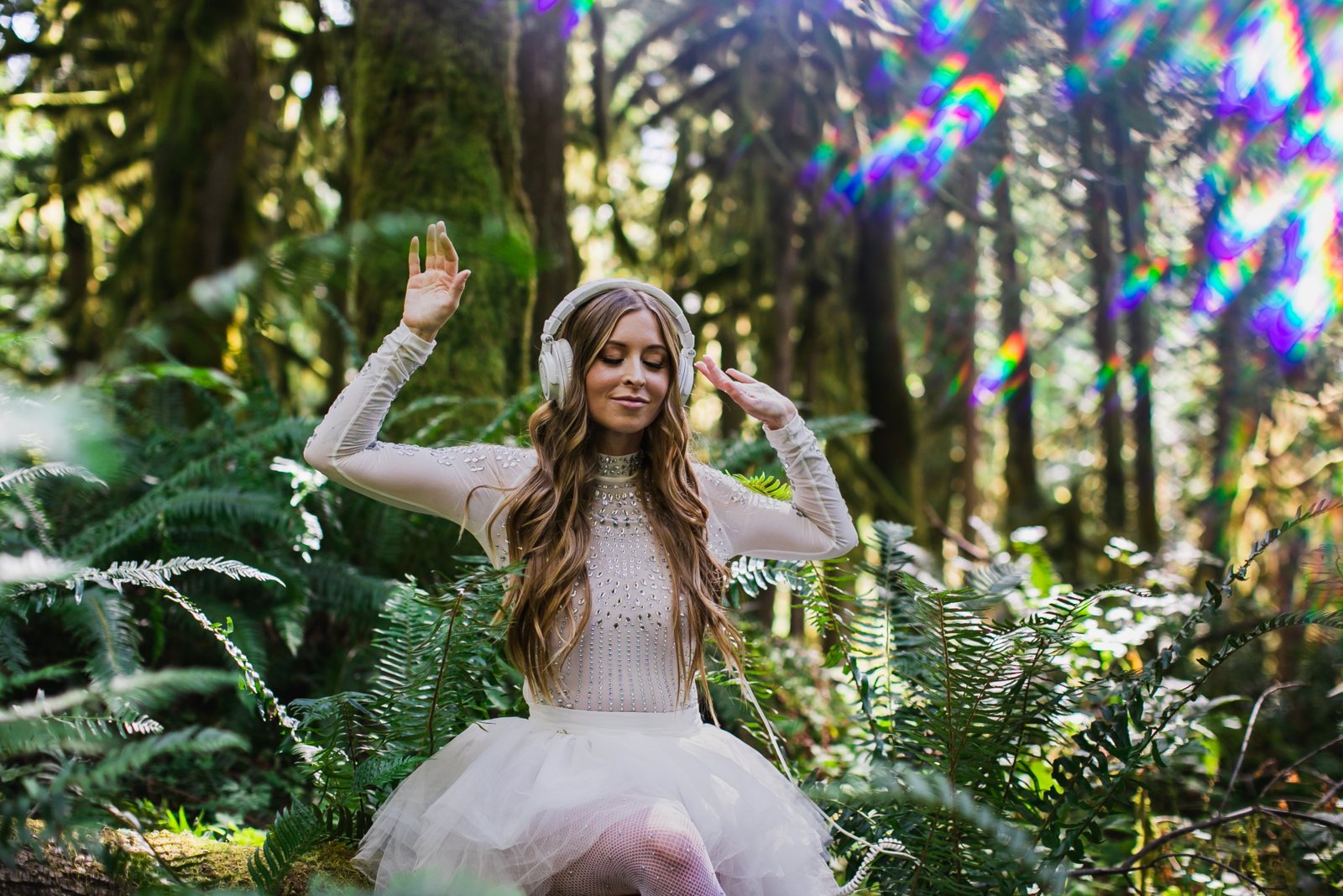 Leanne in white headphones and a crystal-studded bodysuit, eyes closed, dancing with her hands raised among ferns