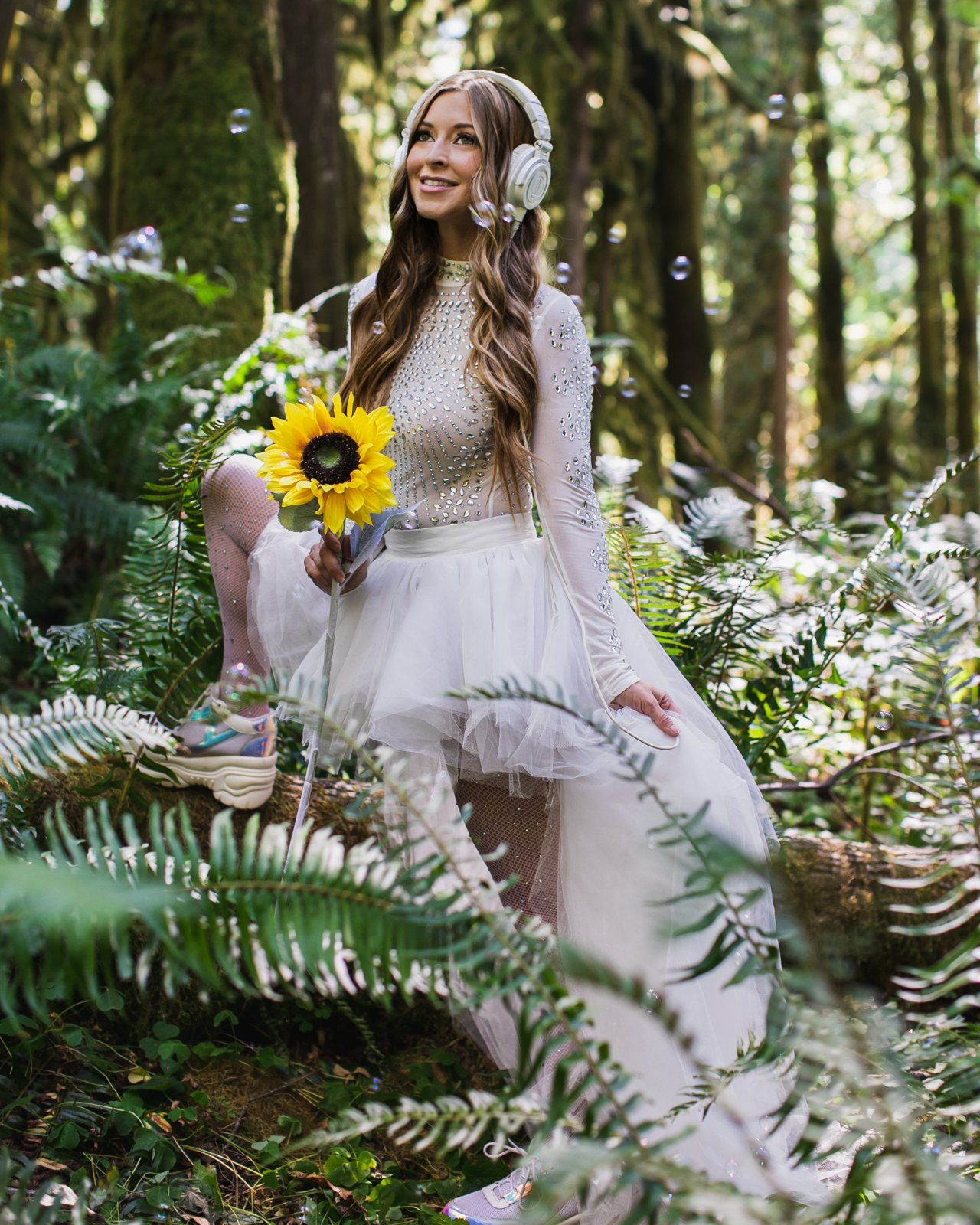 Leanne smiling in white headphones and a tulle skirt, seated on a mossy log holding a sunflower among ferns and bubbles