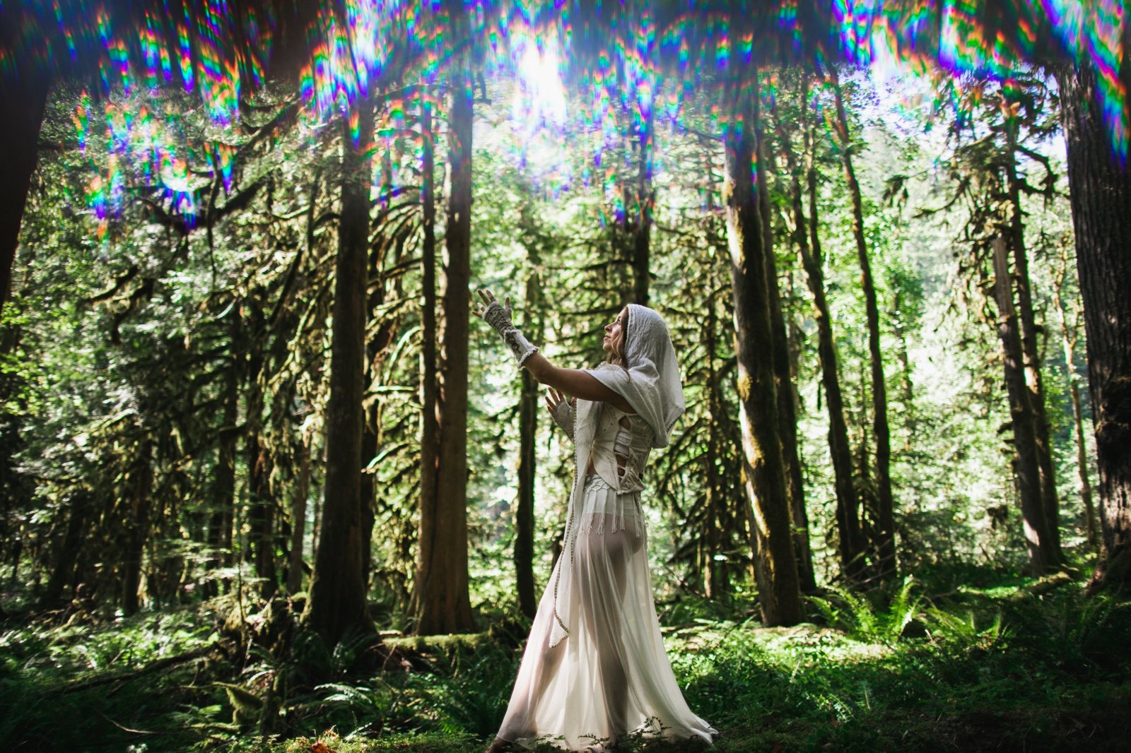 Leanne in a white hooded robe reaching up toward rainbow light falling through a towering mossy forest