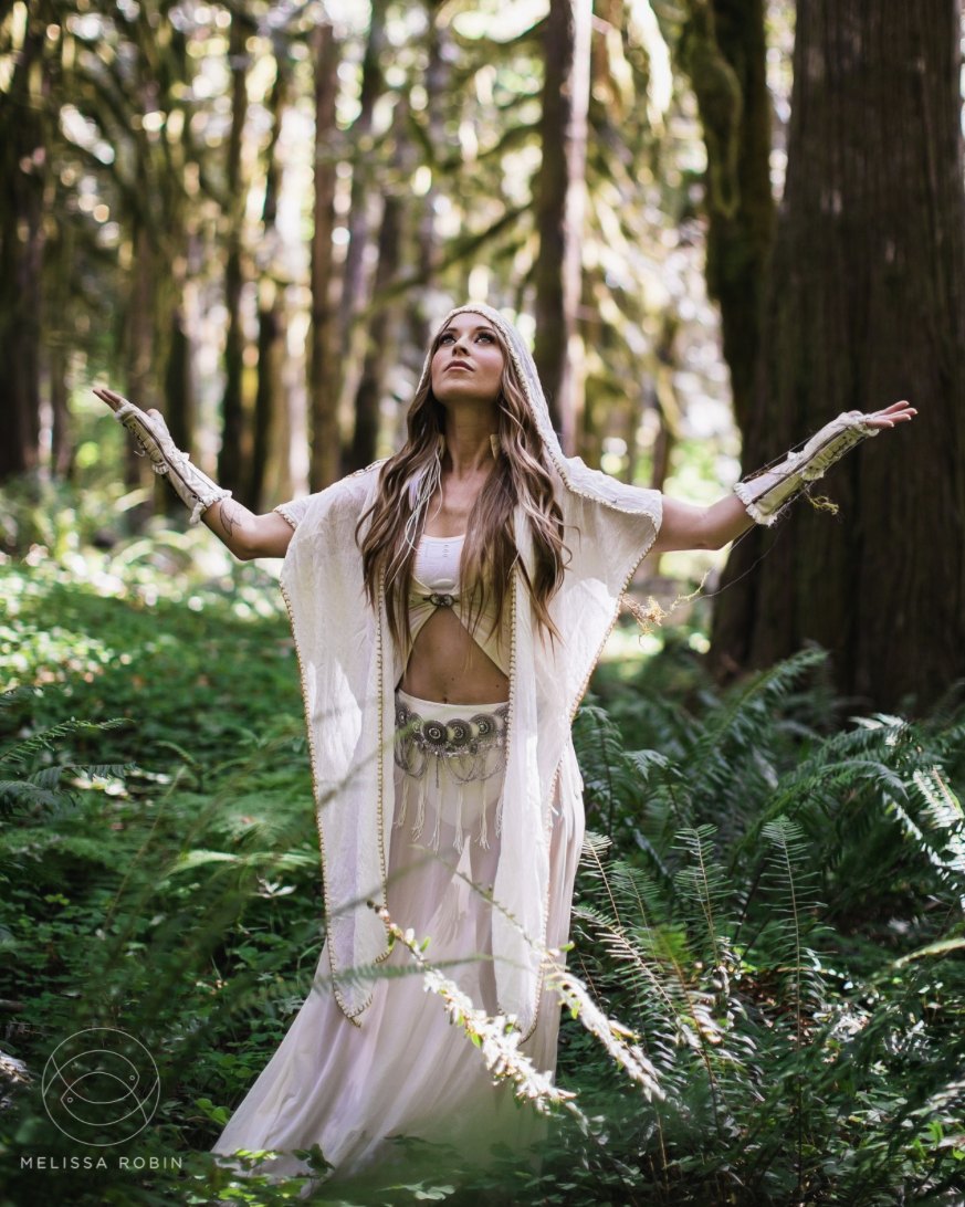 Leanne in flowing white robes and a hood, arms raised to the sky among ferns and tall forest trees