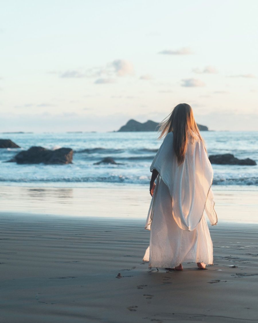 Leanne seen from behind in a flowing white robe, walking toward the ocean at golden hour