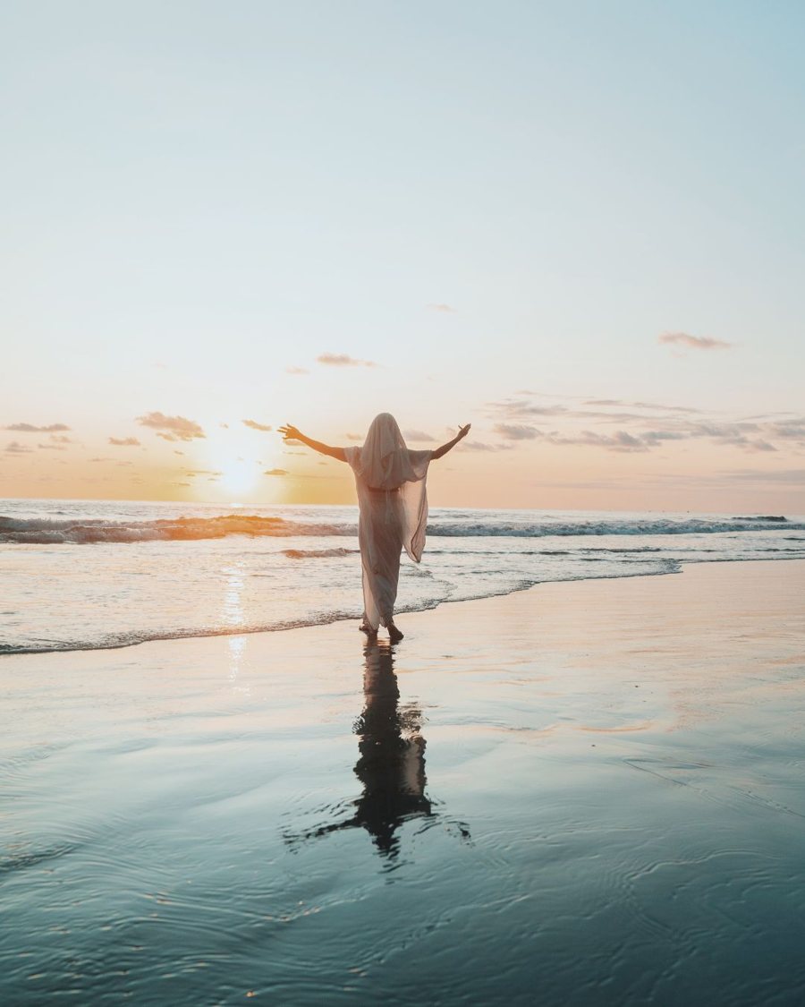 Leanne veiled in white with arms open to the sunset, reflected in the wet sand