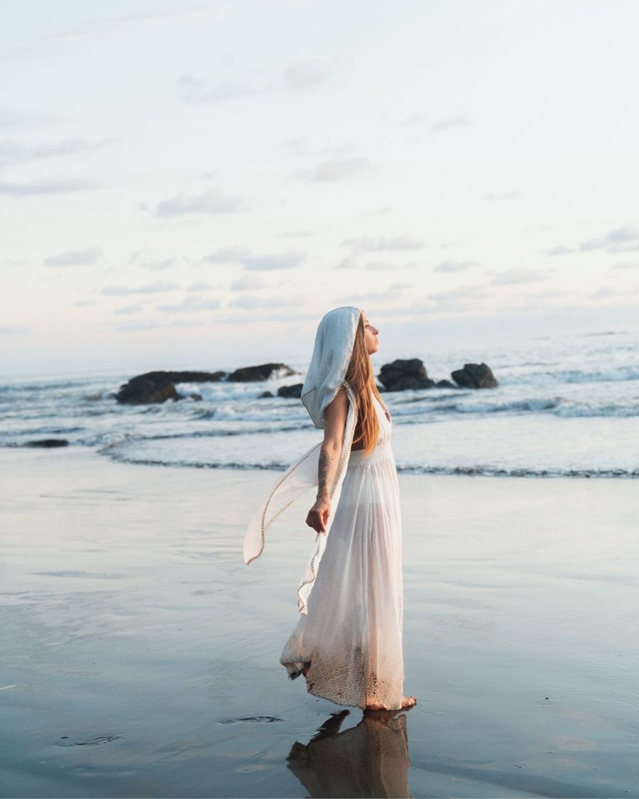 Leanne in a white hooded dress standing at the shoreline, face lifted to the sky
