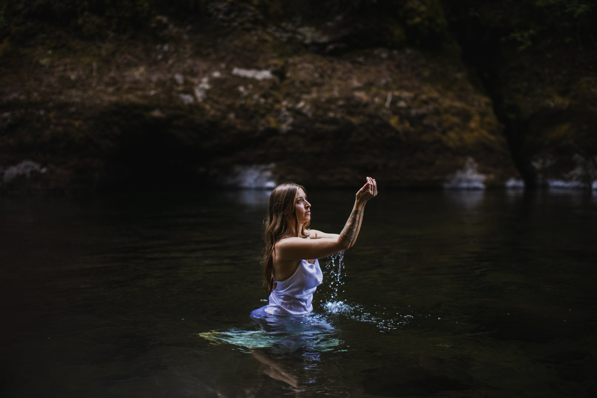 Leanne standing waist-deep in a dark forest pool in a white slip, lifting cupped hands as water streams through her fingers