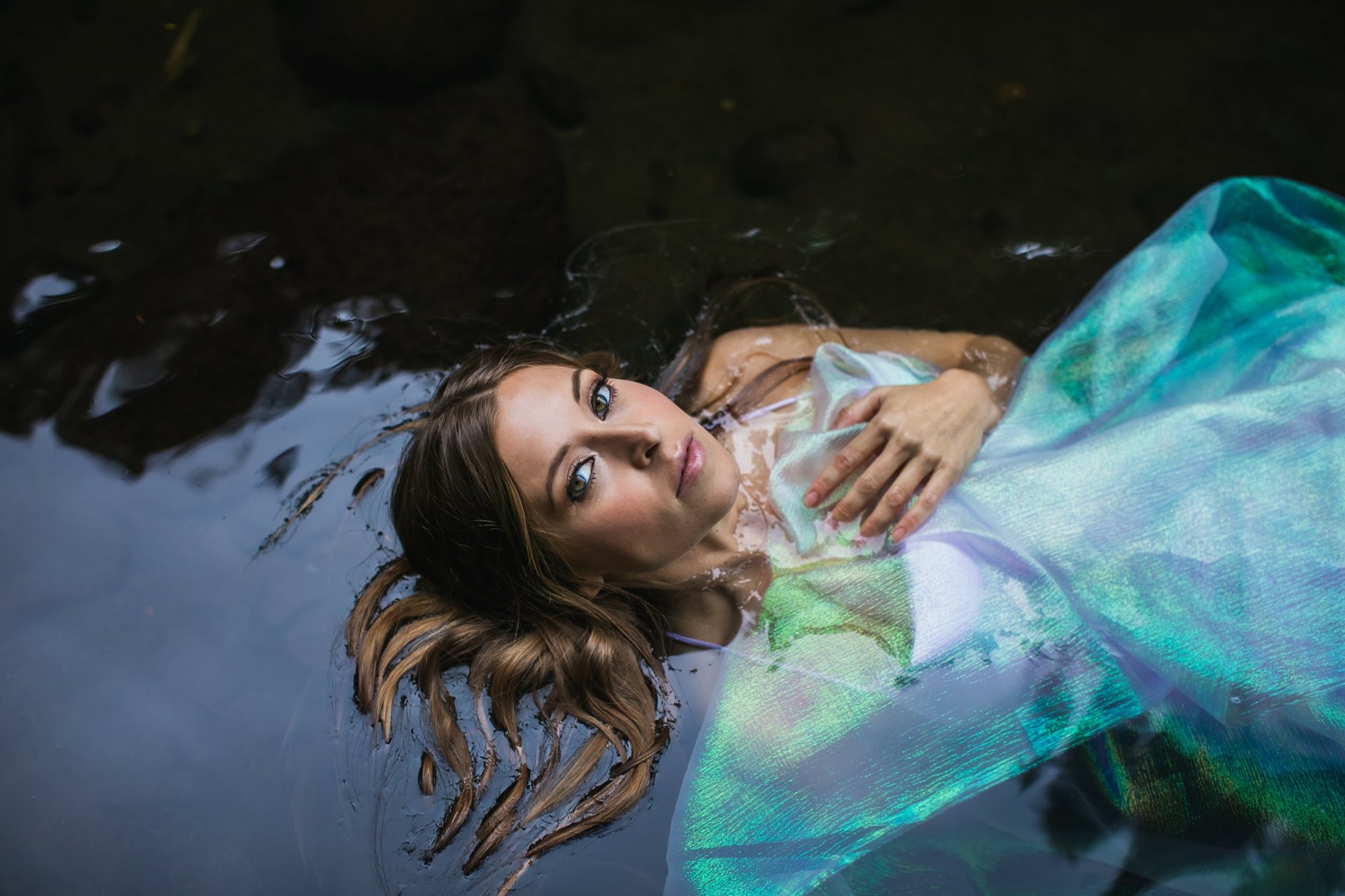Leanne floating on her back in dark water, wrapped in iridescent blue and green fabric, one hand on her heart, gazing up