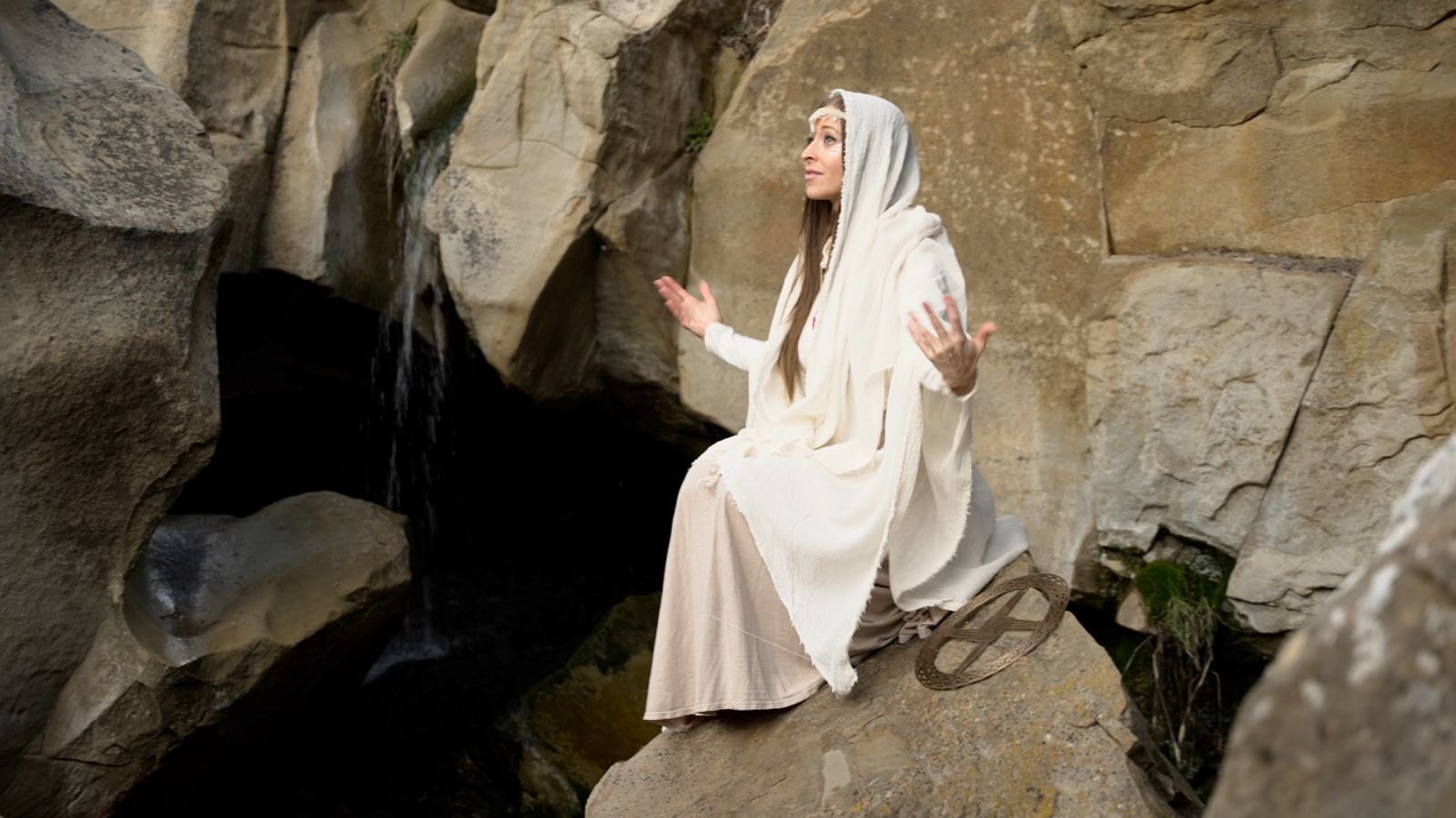 Leanne Falco in flowing white robes, seated on a rock beside a waterfall, arms open in prayer