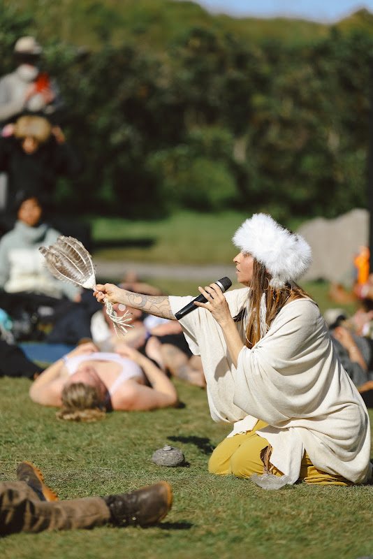 Leanne kneeling on the grass singing into a microphone and fanning a feather over a resting crowd