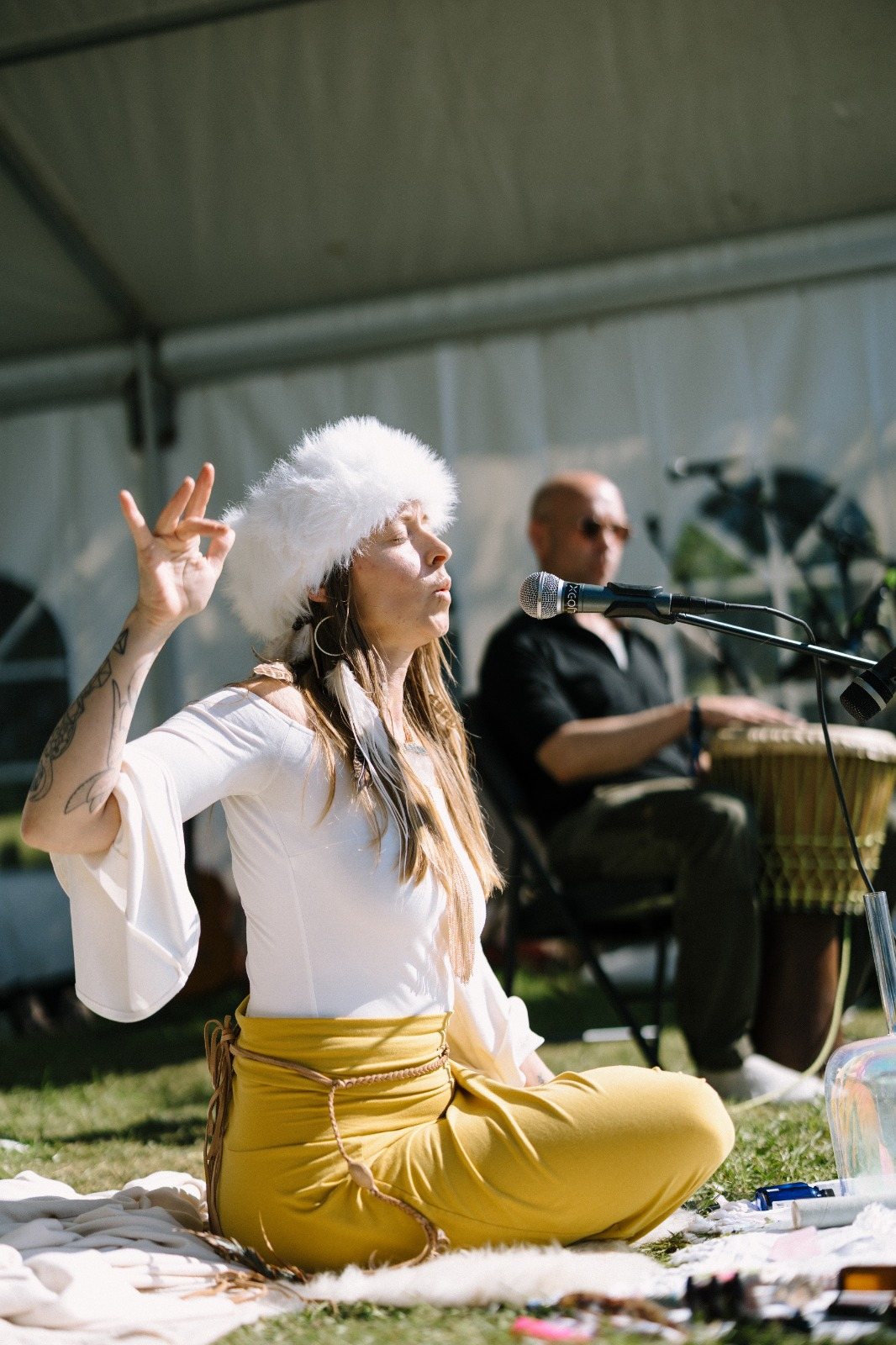Leanne seated cross-legged in white and mustard, eyes closed, singing into a microphone with a drummer behind her