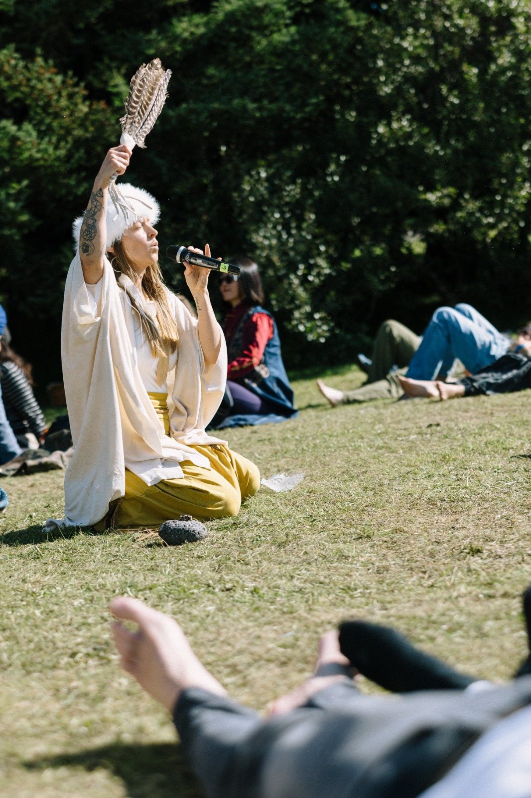 Leanne kneeling in the sun, feather fan raised high, singing into a microphone