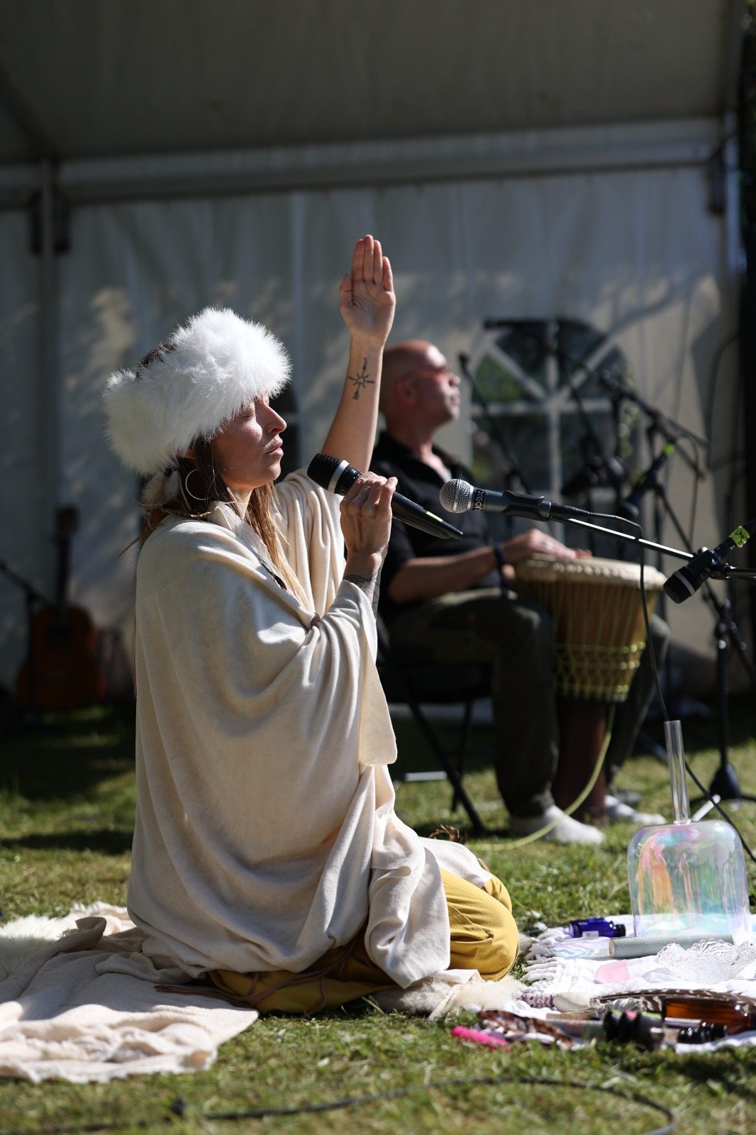 Leanne in a white fur hat, eyes closed, one hand raised as she sings under the tent with a drummer behind her