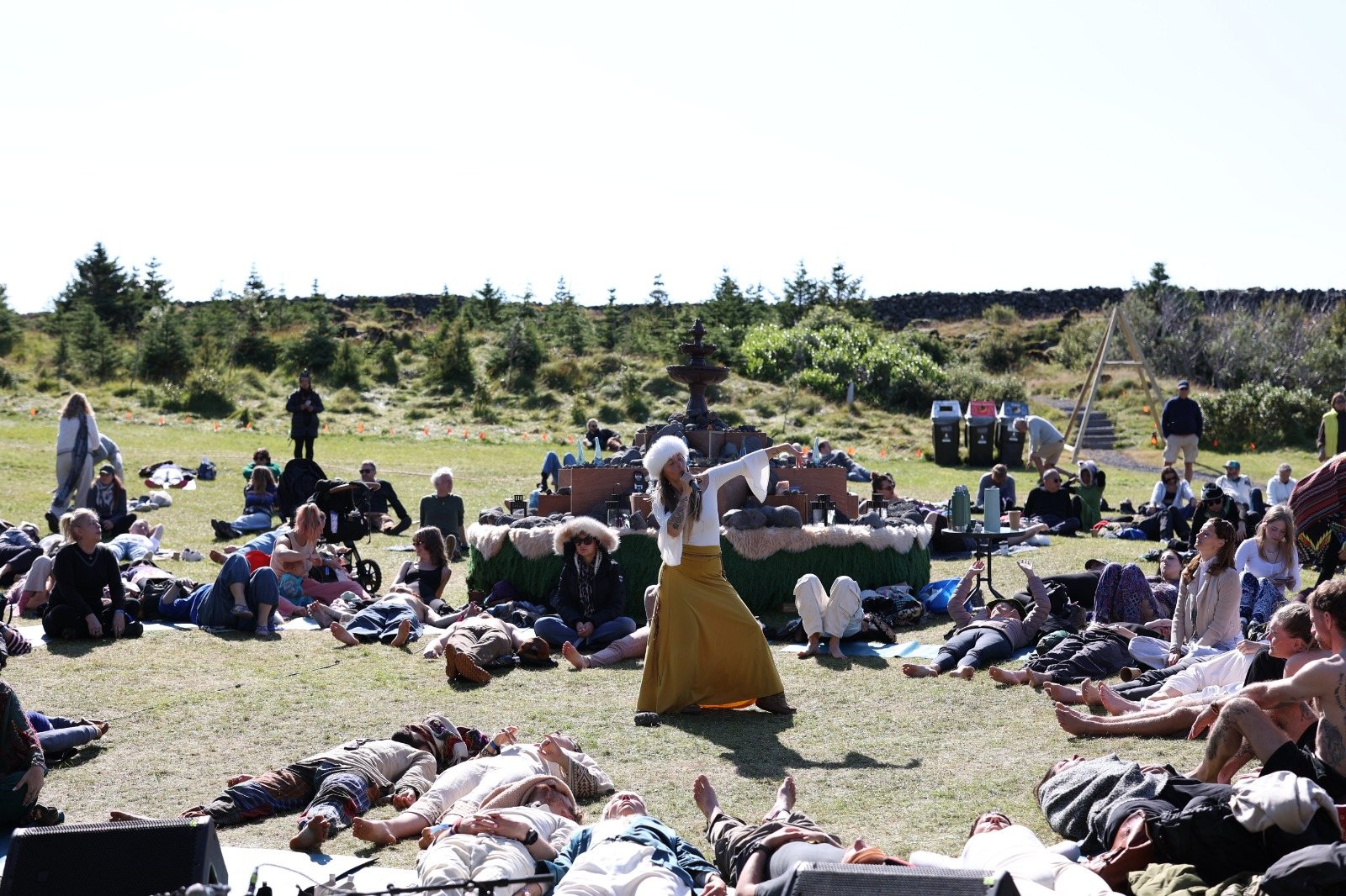 Leanne in a white fur hat and mustard skirt singing among a field of people resting on the grass