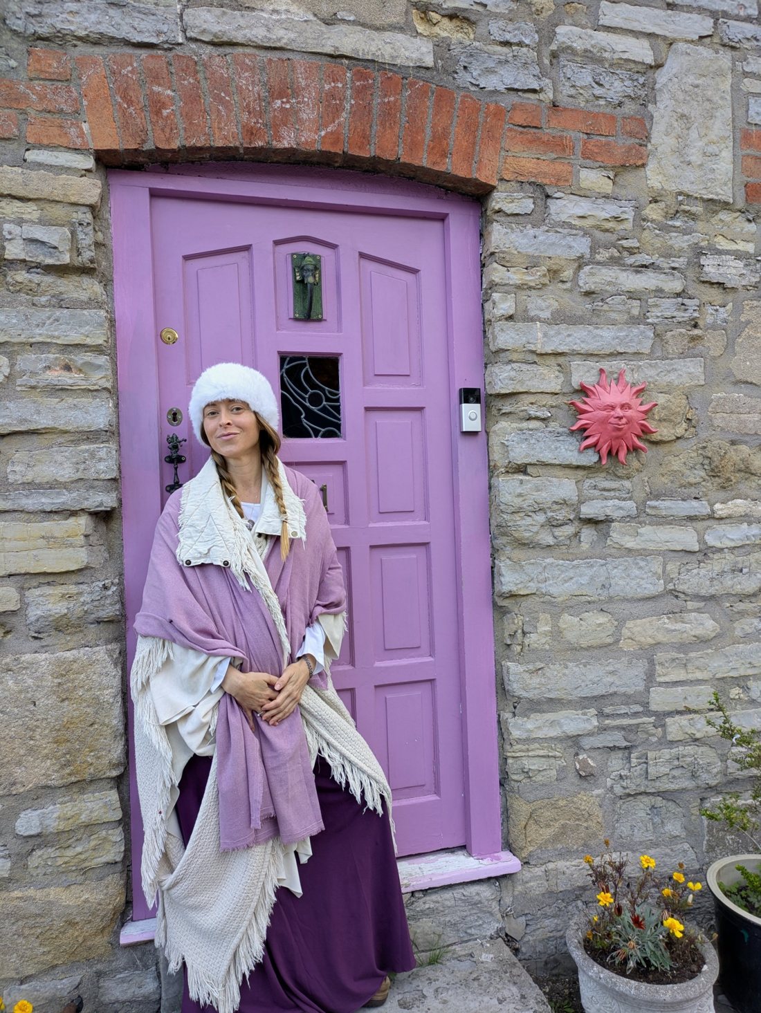 Leanne in a white fur hat and lavender shawls at a lilac door in an old stone wall