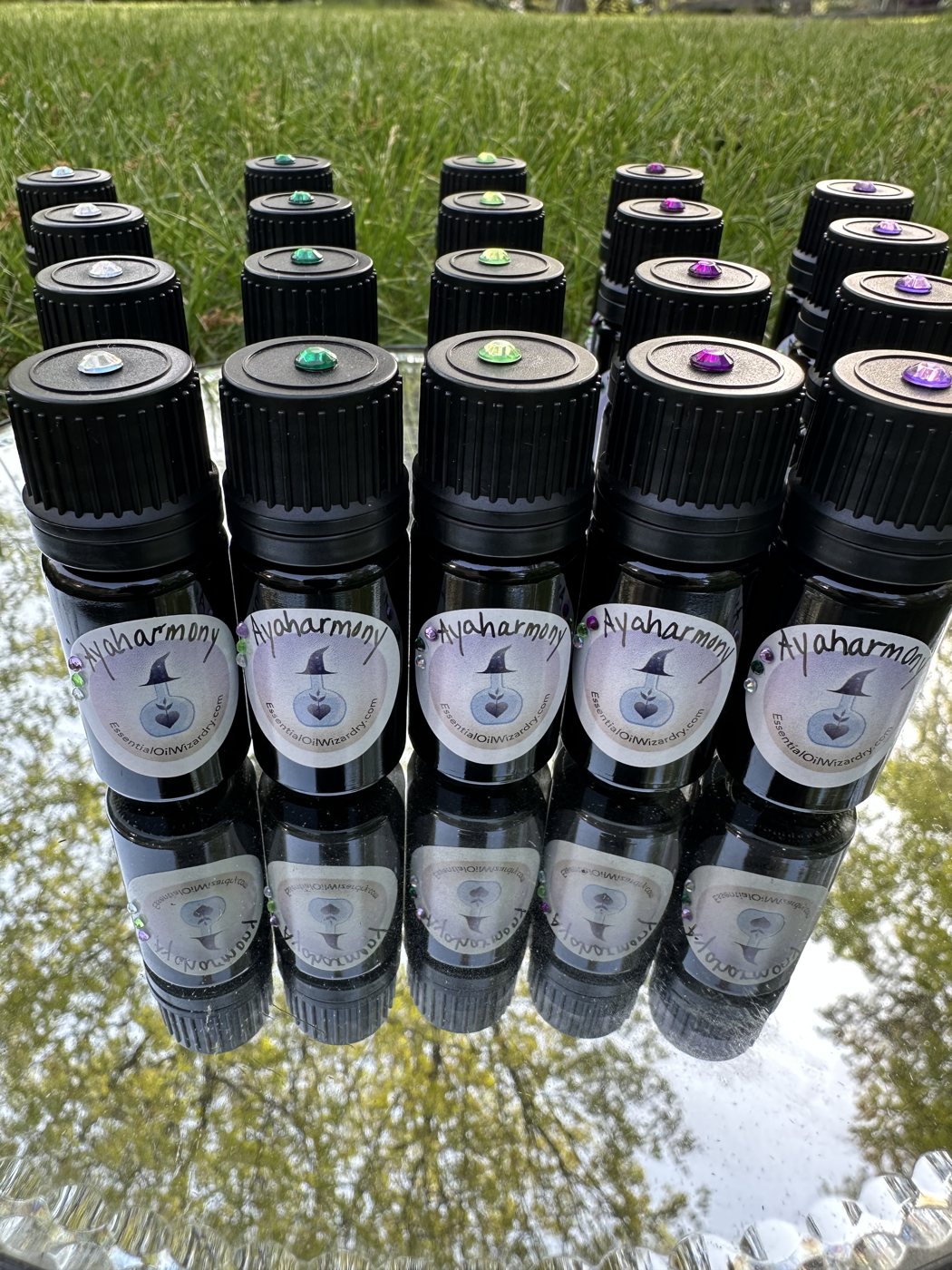 Rows of small dark Ayaharmony essential oil bottles with crystal-topped caps, reflected on a mirror tray in the grass