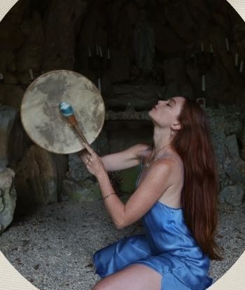 Fiire Love kneeling in a stone grotto, playing a frame drum