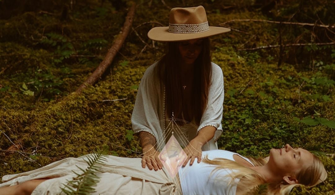 Leanne in a wide-brimmed hat with her hands resting on a woman lying on the moss in a forest