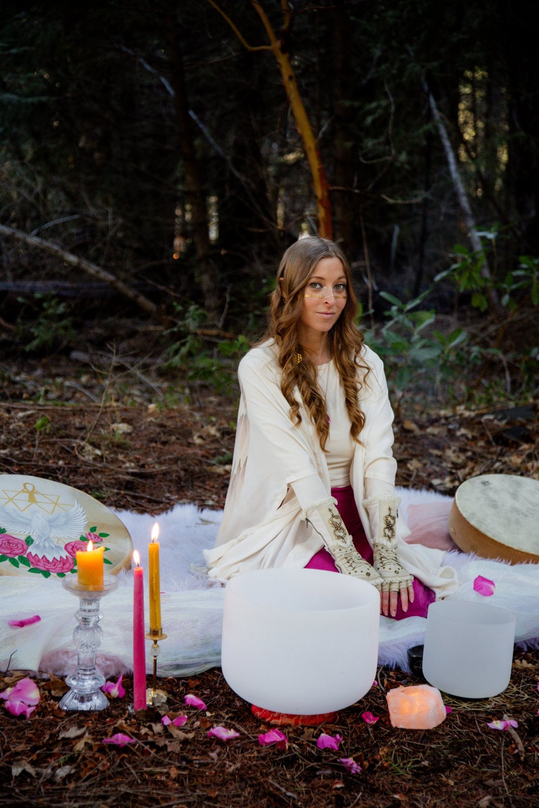 Leanne seated in the forest among crystal singing bowls, frame drums, candles and rose petals