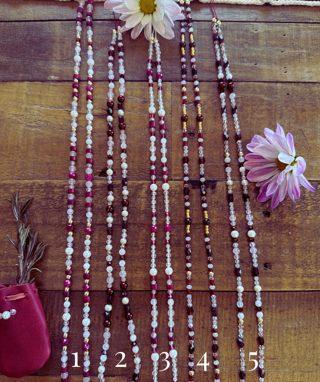 Five strands of womb beads laid side by side on a wooden table, numbered 1 to 5, with a red leather pouch, rosemary and pink flowers